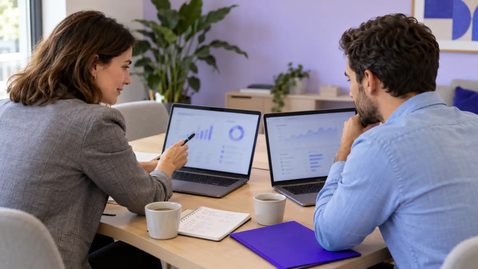 Dos profesionales comparan en sus portátiles dos paneles de CRM en una oficina, con una libreta de cálculos sobre la mesa de reuniones
